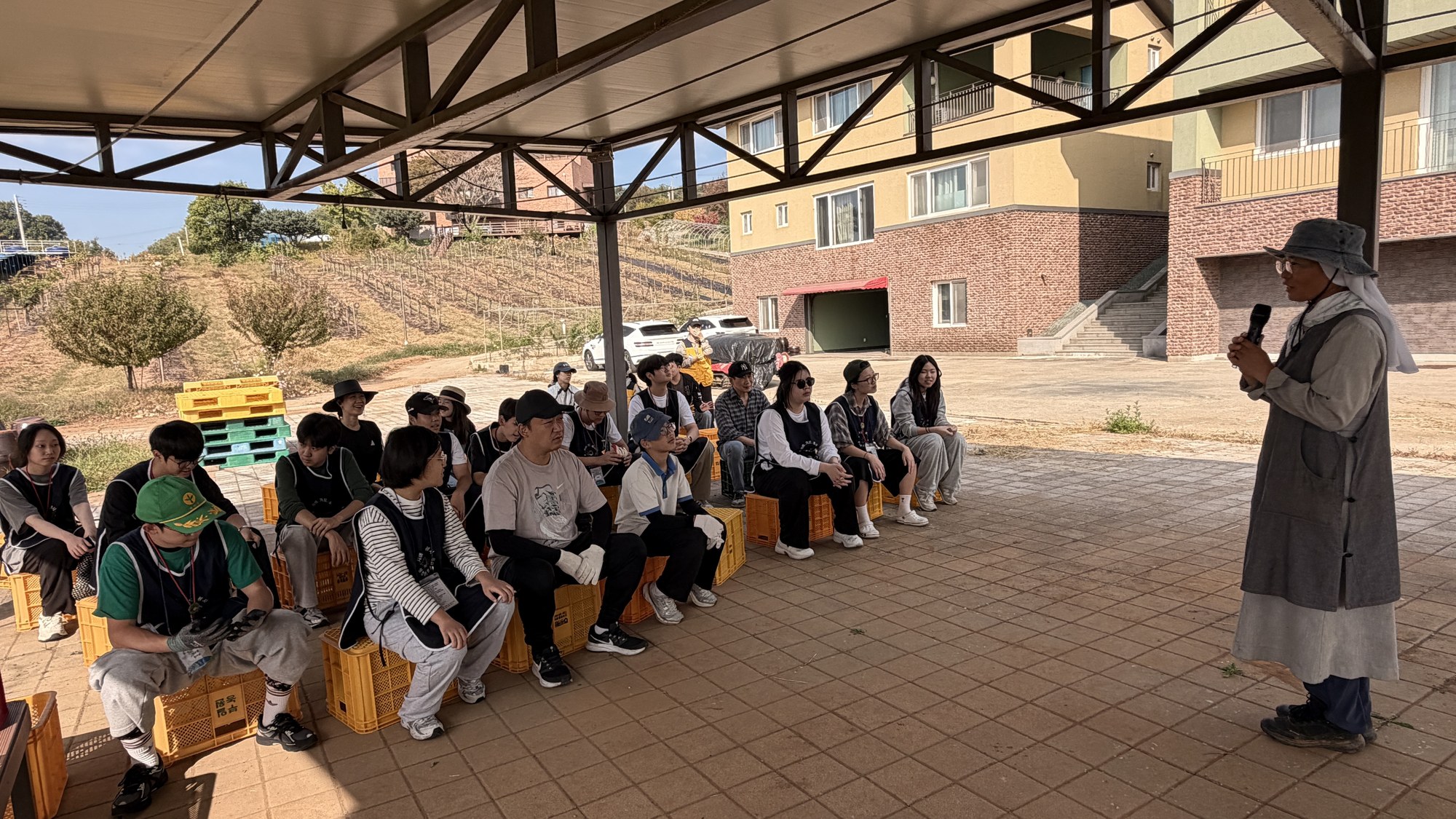 지붕 아래 노란 상자에 앉아 마이크를 든 수녀님의 이야기를 듣는 청소년들