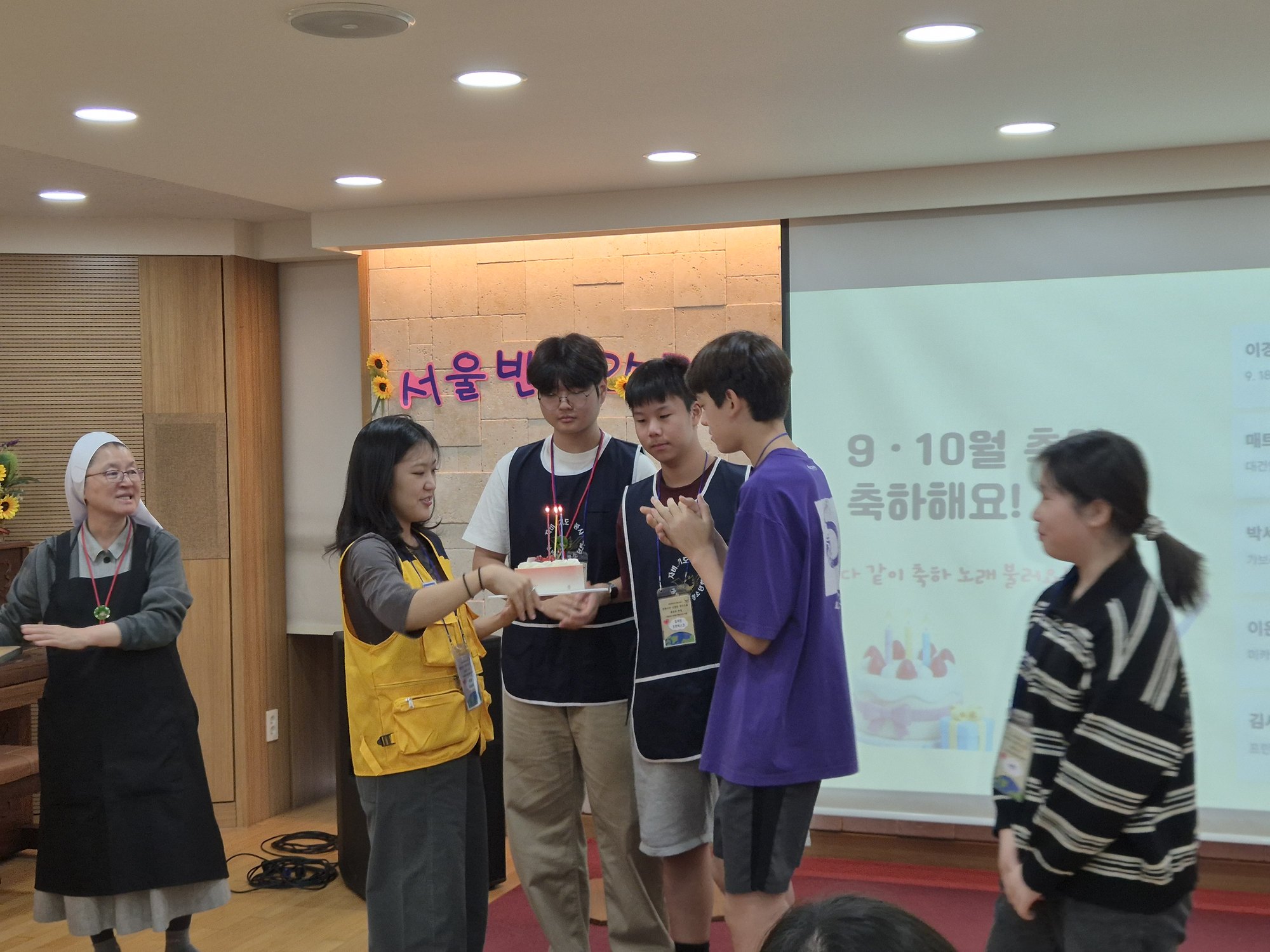 ‘9 · 10월 축일 축하해요!’ 화면 앞에서 케이크를 받는 친구들과 웃으며 지켜보는 수녀님