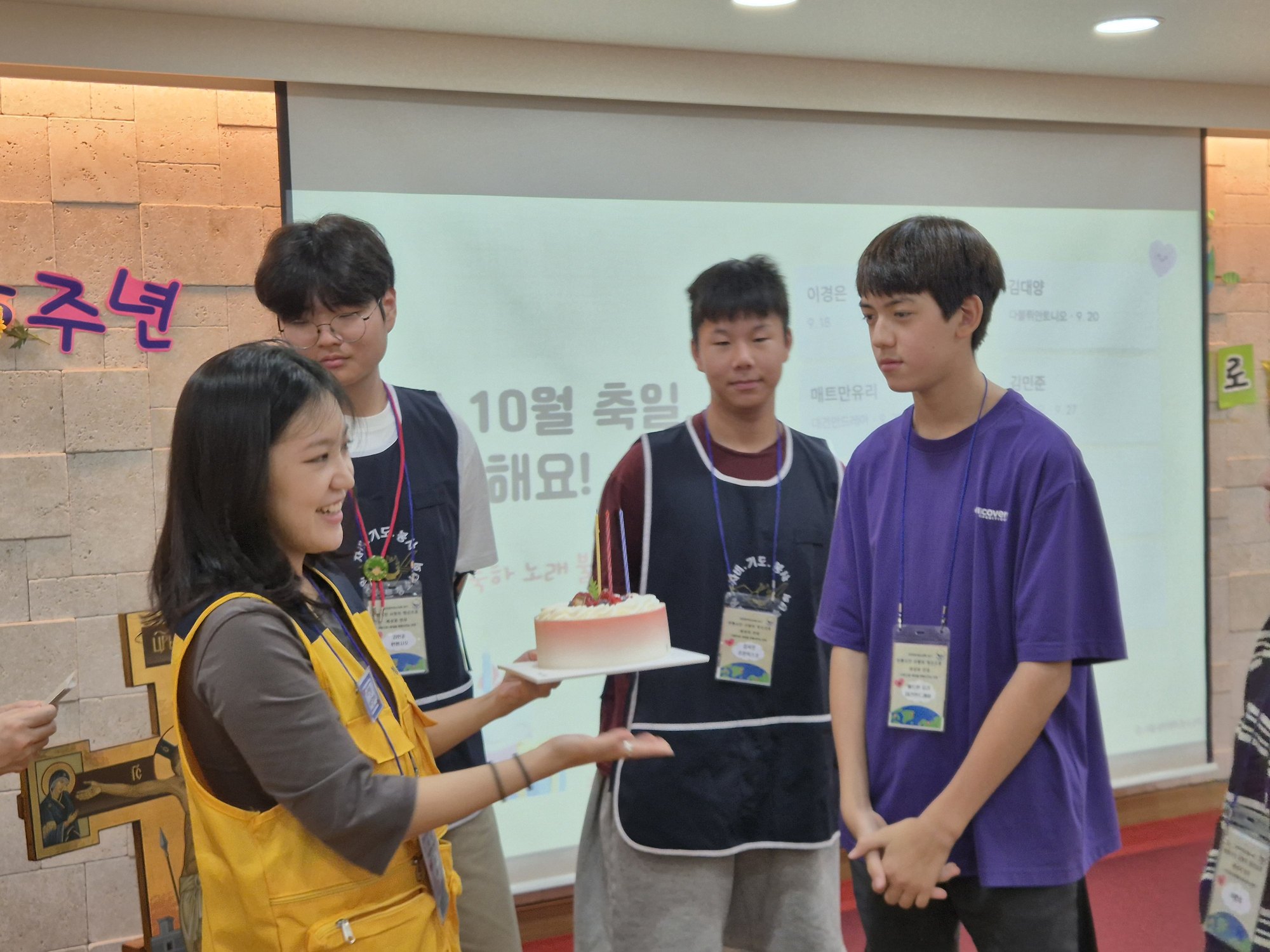 노란 조끼를 입은 선생님이 활짝 웃으며 축일 친구들에게 케이크를 내미는 모습